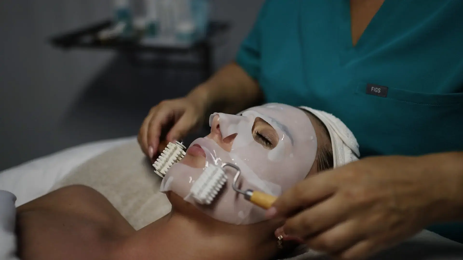 A practitioner applies a sheet mask to a client's face with a facial roller