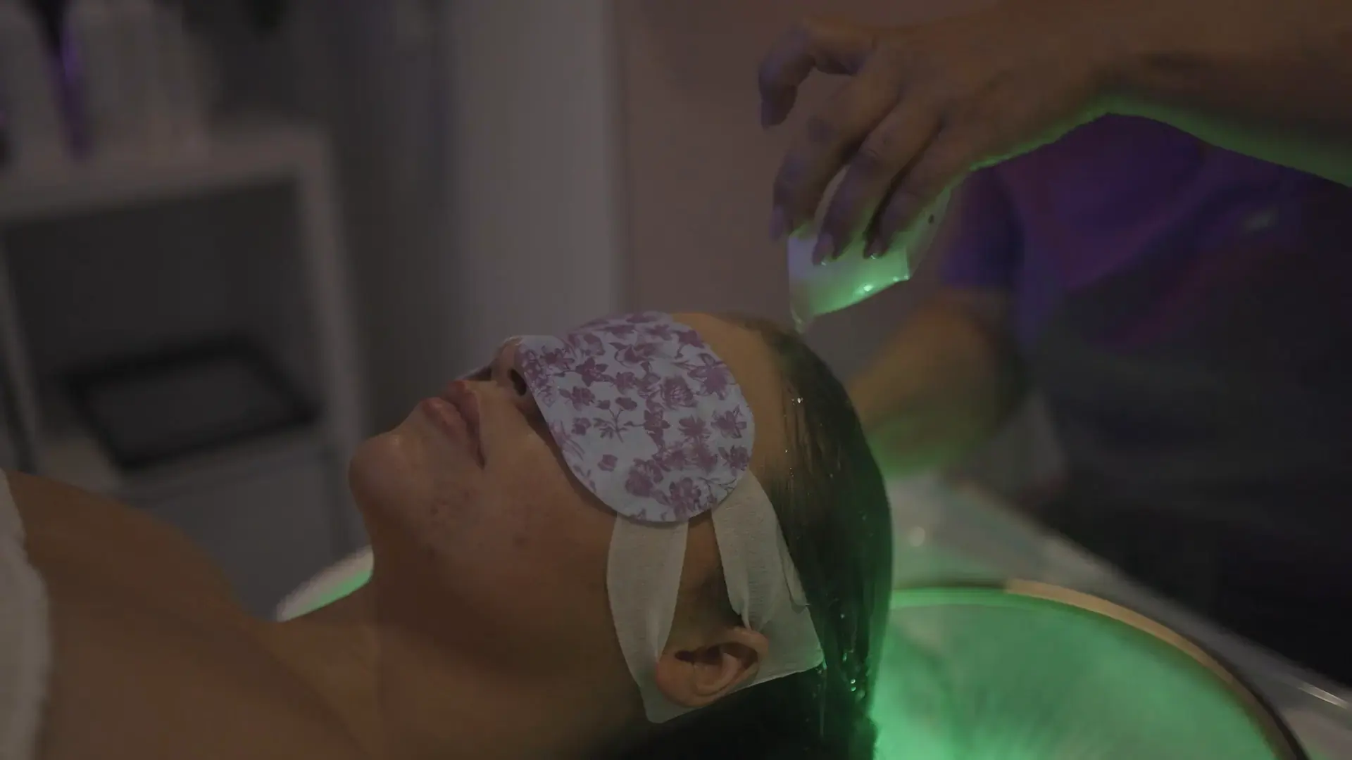 A client wearing a patterned eye mask lies in soft green light during a head spa treatment