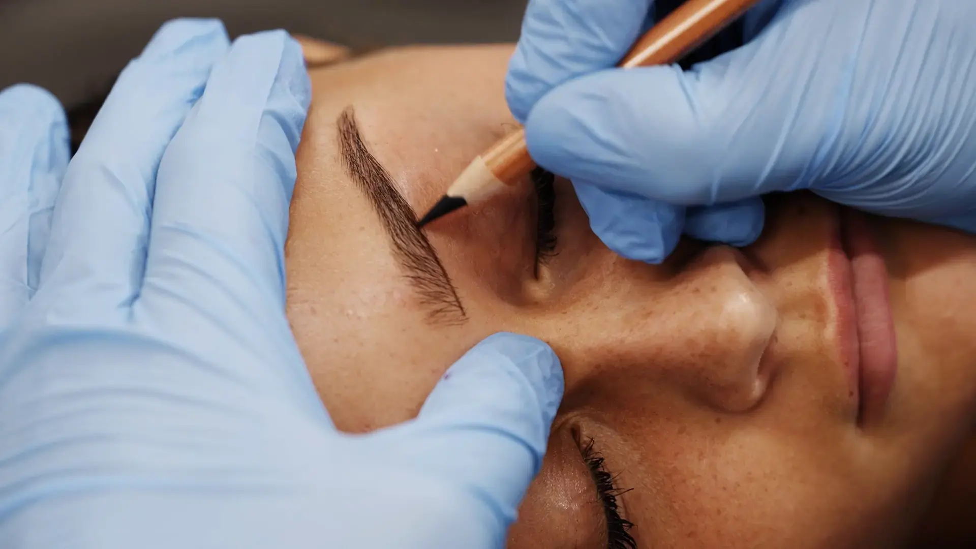 A practitioner draws a brow shape in pencil on a client's face with gloved hands