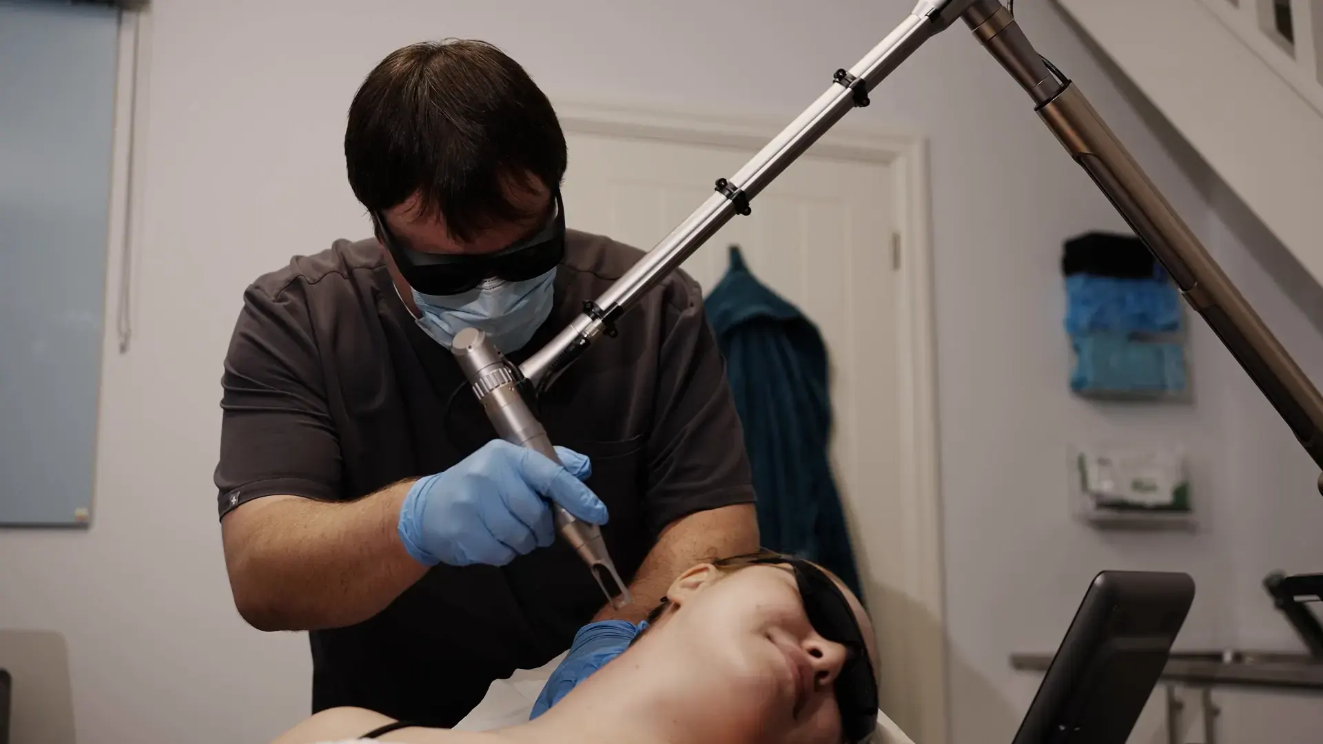 A practitioner in protective glasses uses a laser handpiece on a client during tattoo removal