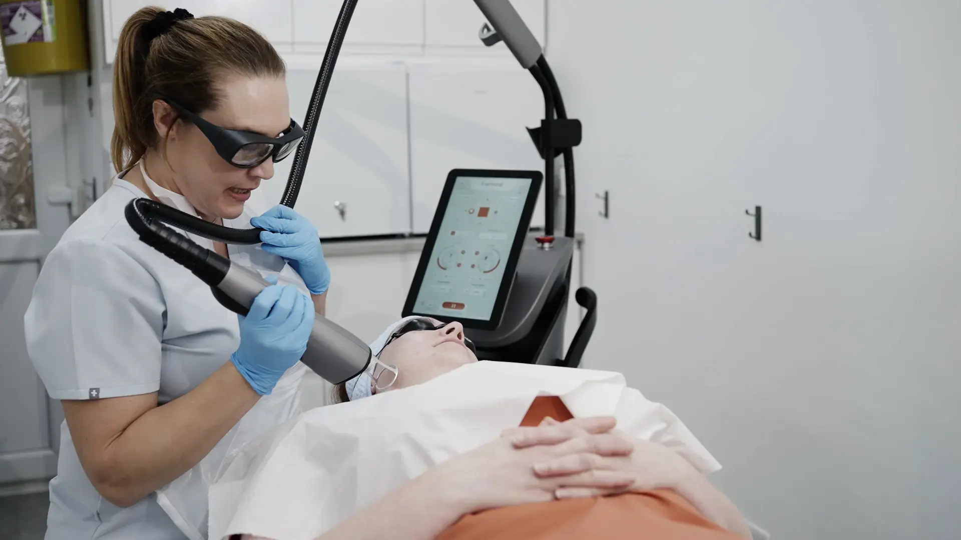 A practitioner in protective glasses holds a laser handpiece over a client lying on a couch, with a touchscreen machine beside her