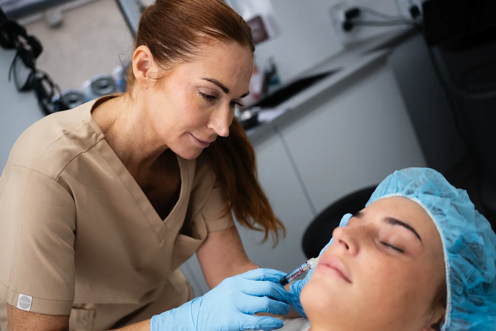 A practitioner in a beige tunic holds a syringe near a client's cheek