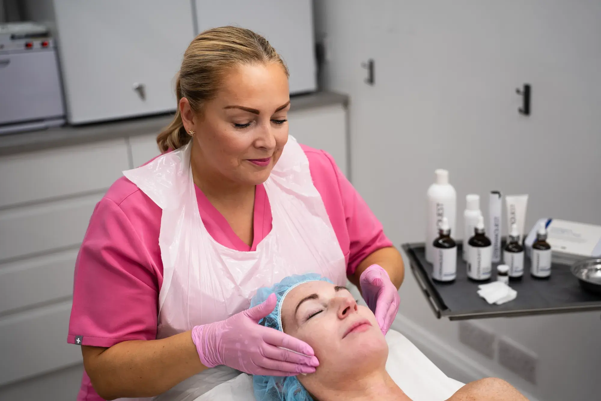 A practitioner in a pink tunic treats a client's face, with skincare products on a trolley beside them