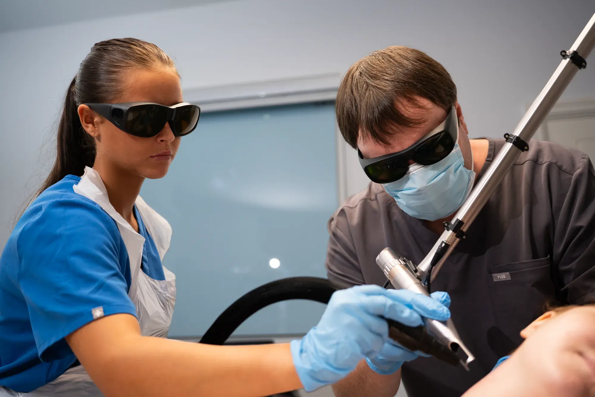 Two practitioners in protective glasses use a laser handpiece on a client during tattoo removal