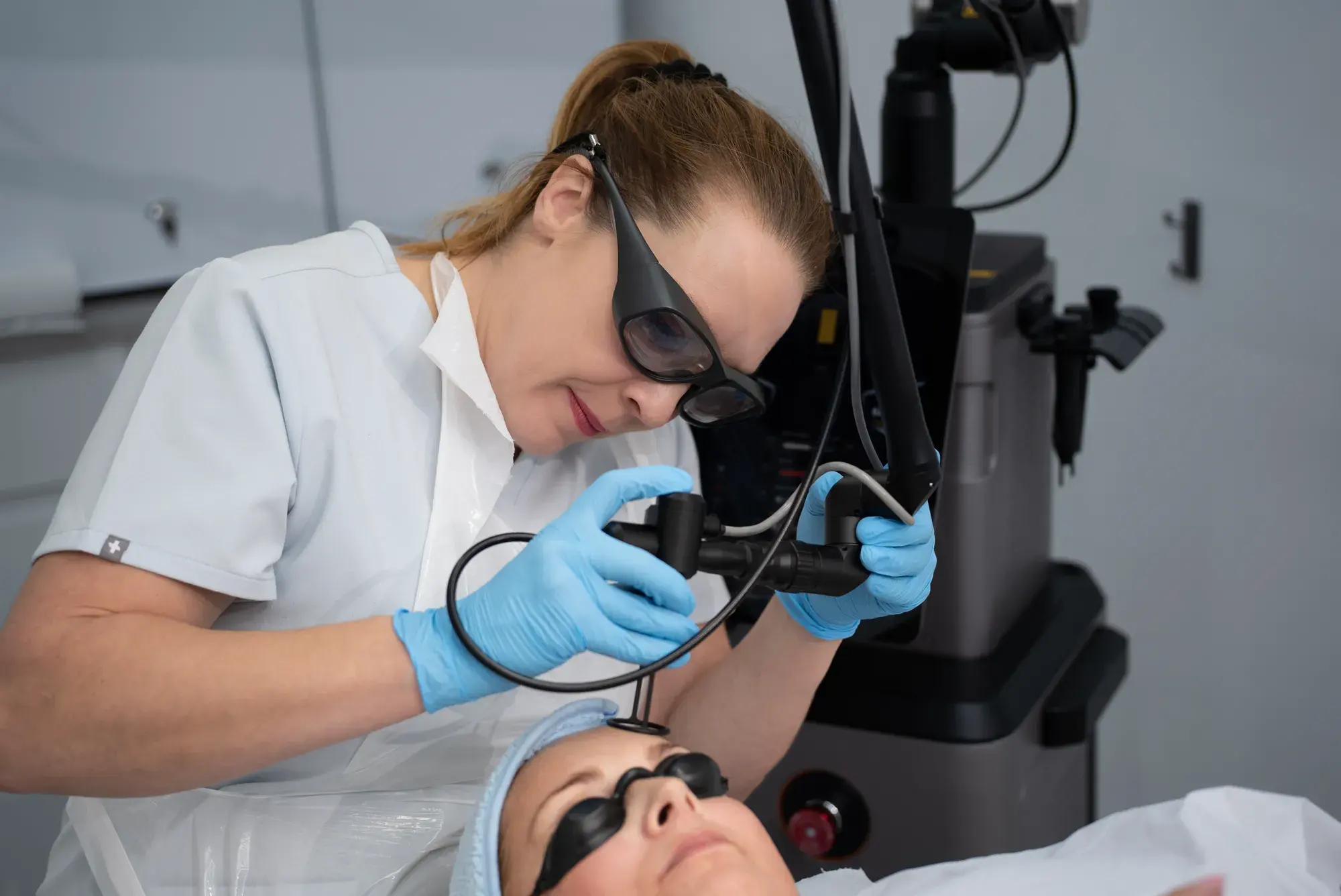 A practitioner in protective glasses uses a CO2 laser handpiece on a client's face