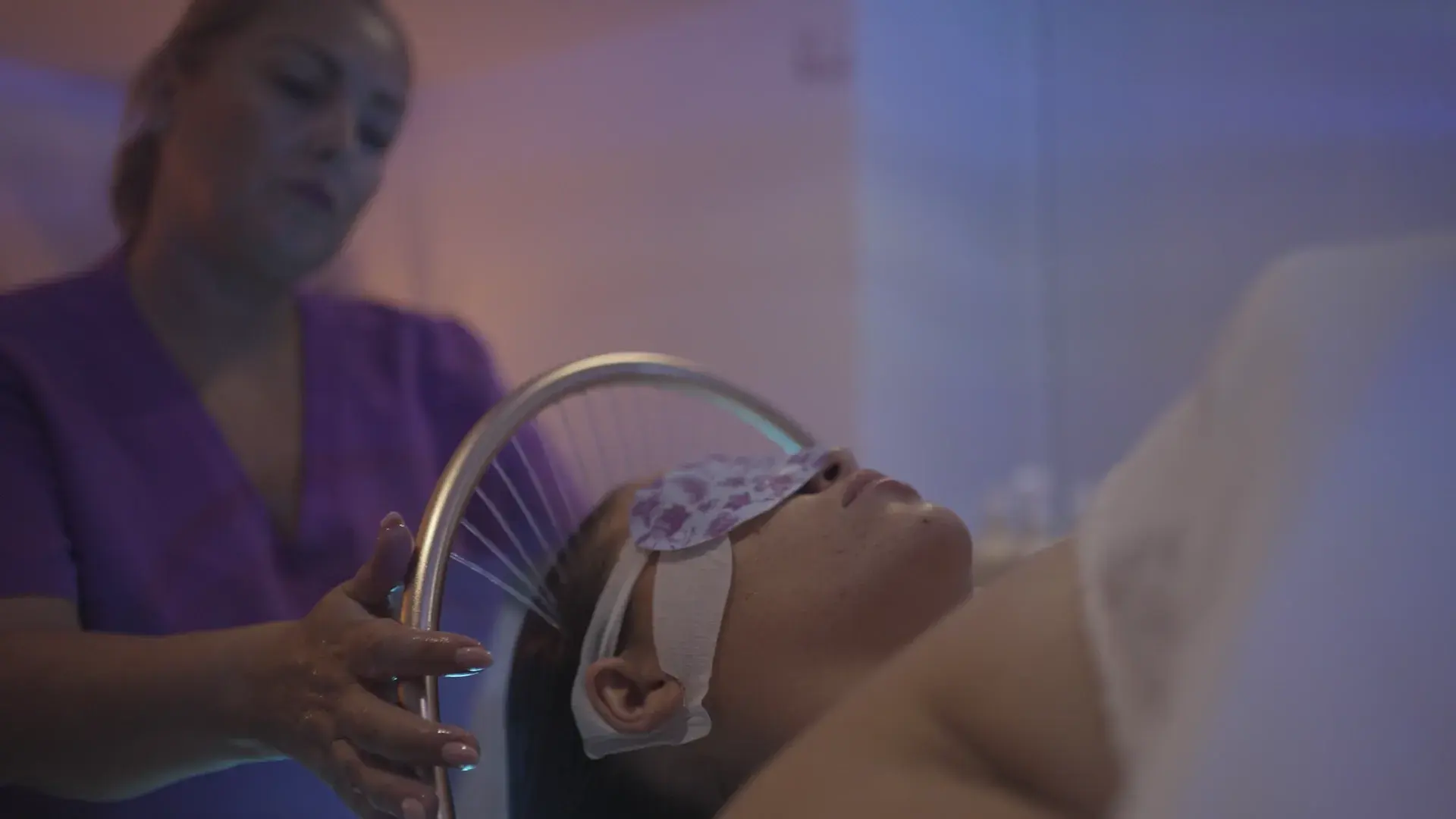 A practitioner in a purple tunic holds a shower head over a client wearing an eye mask in coloured light