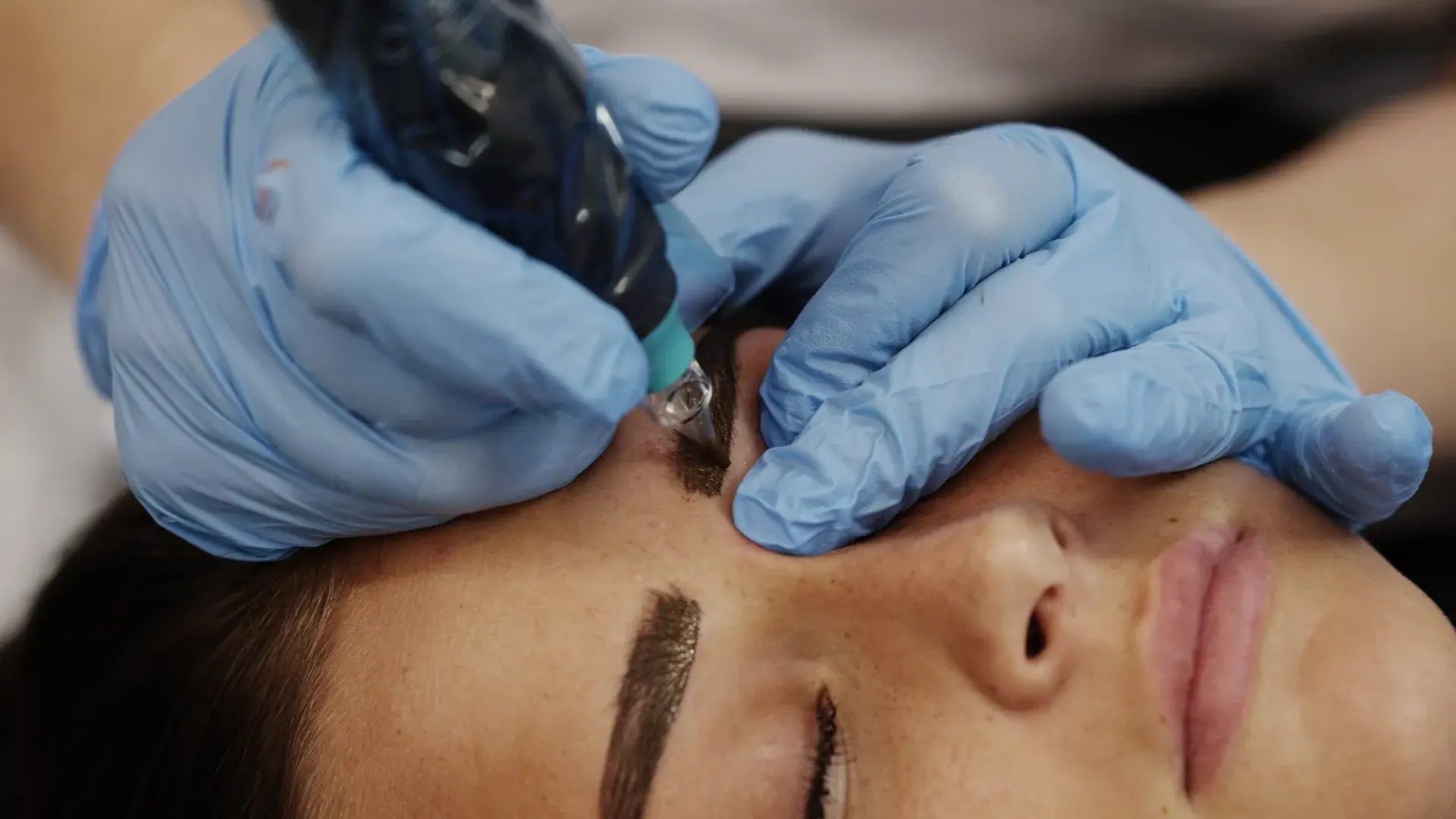 A practitioner holds a tattoo machine handpiece beside a client's brow and eye