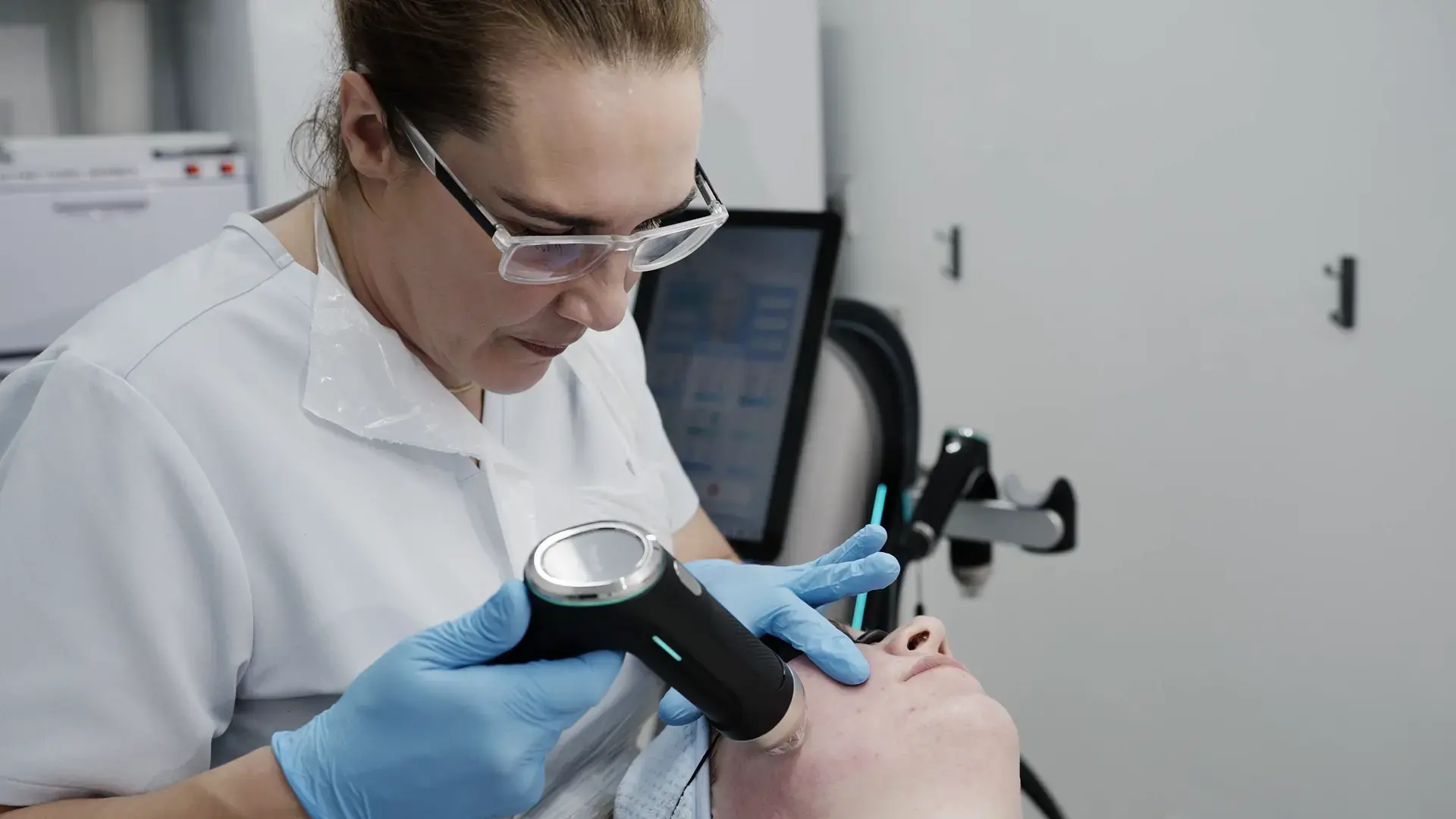 A practitioner in a white tunic holds a radio frequency handpiece to a client's face