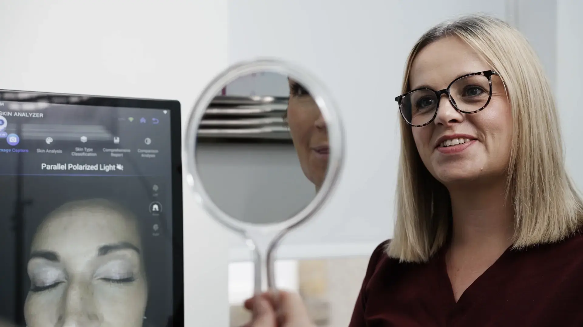 A smiling practitioner in glasses sits beside a skin analysis screen while holding up a hand mirror