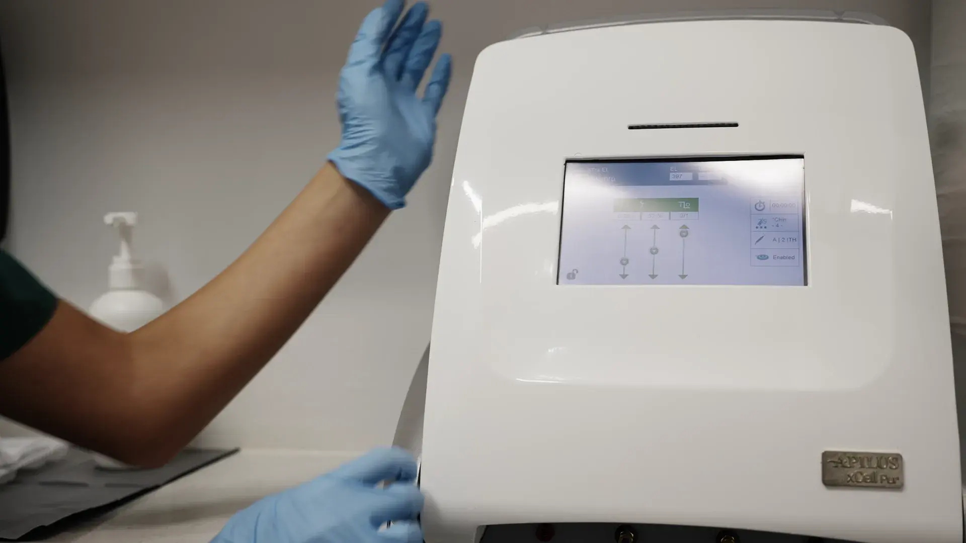 A practitioner in blue gloves stands beside a white electrolysis machine with a touchscreen